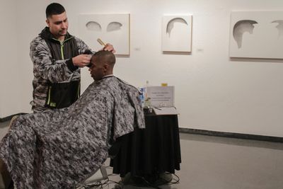 Jose Flores, Barber, cuts hair at opening of Custom Lives, art by Nery Gabriel Lemus in background. Photo by Damian Kelly.